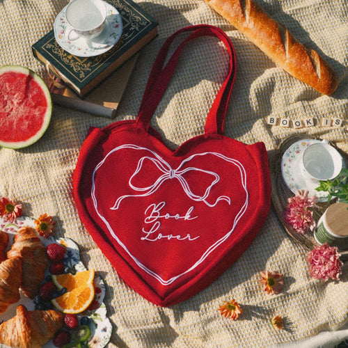 Red heart-shaped bag with 'Book Lover' text on a blanket with food and books.