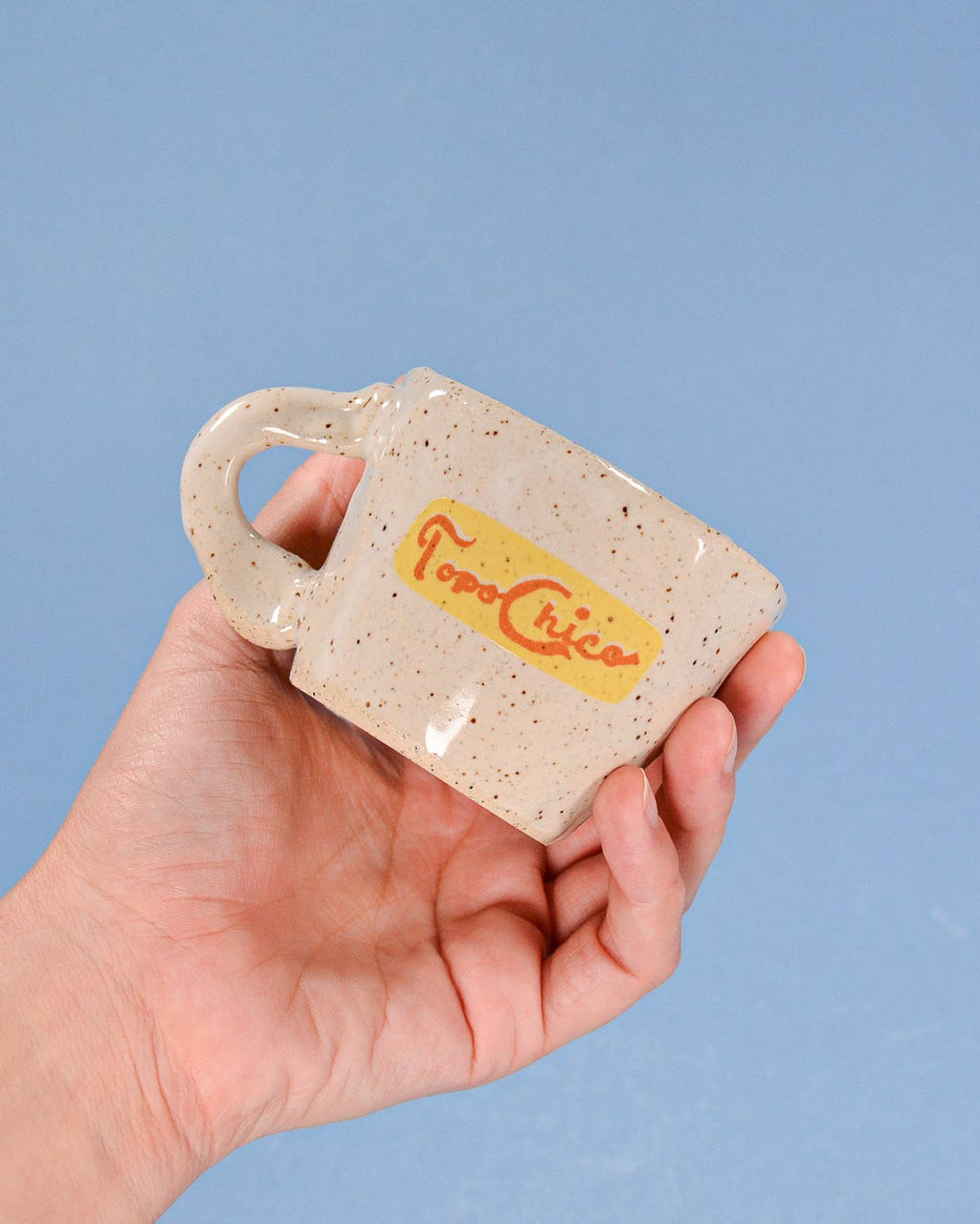Hand holding a small ceramic mug with 'Topo Chico' branding against a blue background