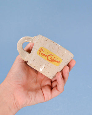 Hand holding a small ceramic mug with 'Topo Chico' branding against a blue background