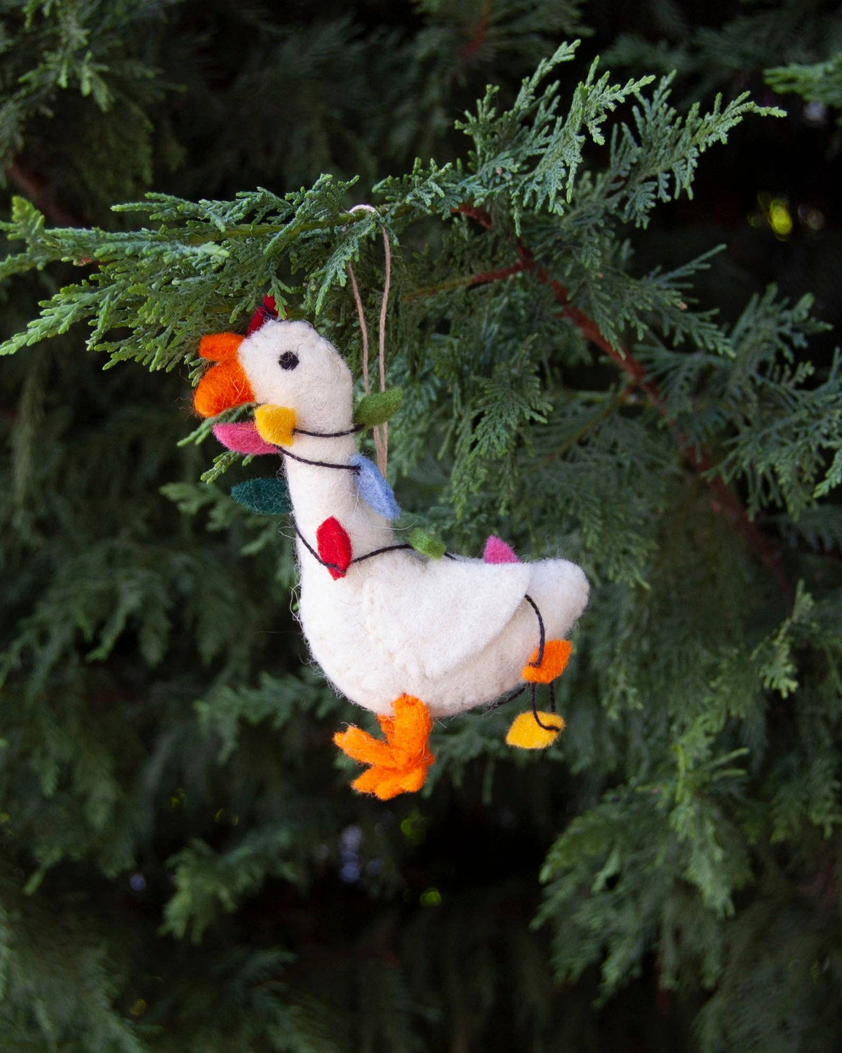 A handmade white felt goose ornament with orange beak and feet, a red comb, and a blue scarf with red hearts, hanging from a string on a green evergreen tree branch.