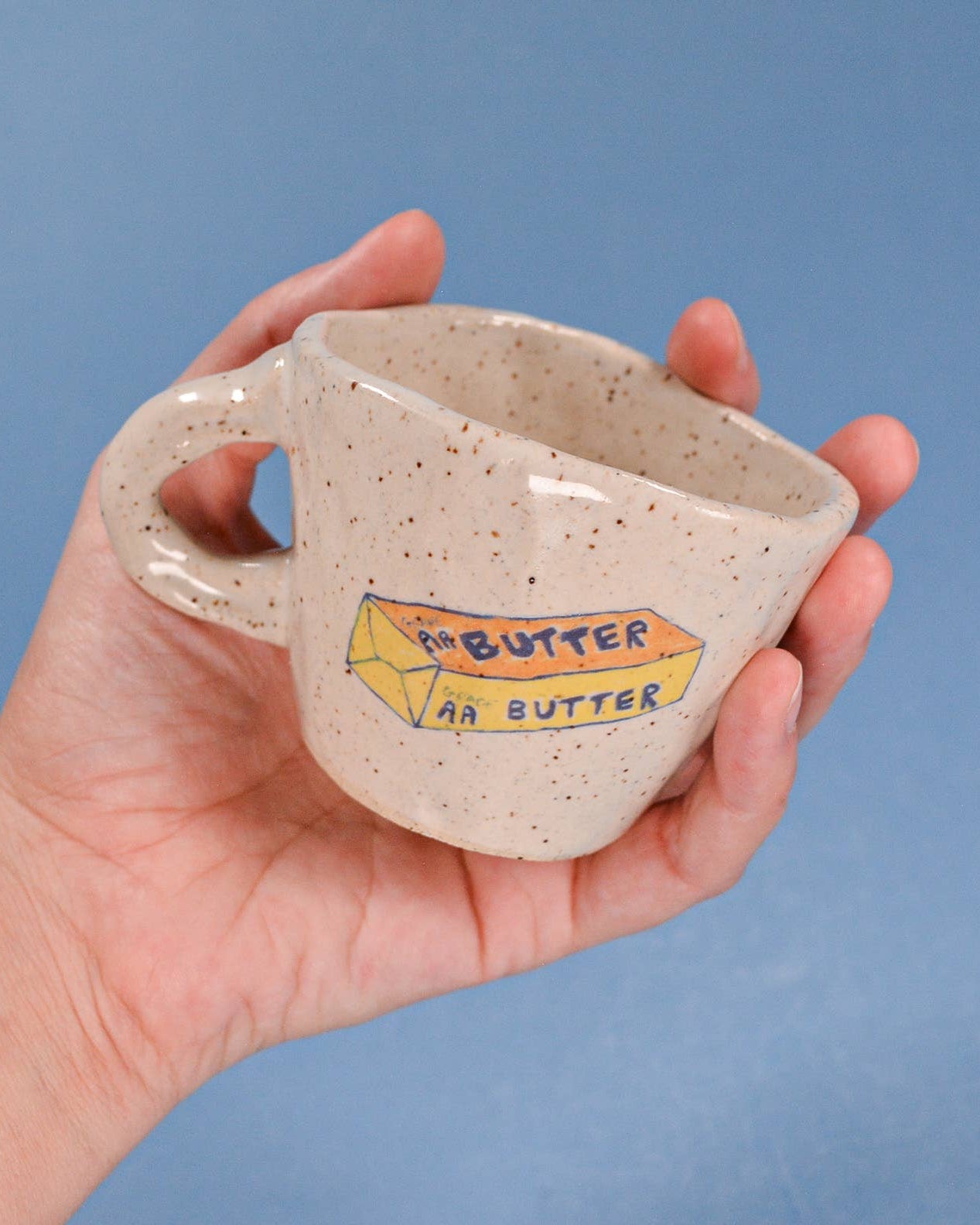 Hand holding a ceramic mug with 'Peanut Butter' label against a blue background