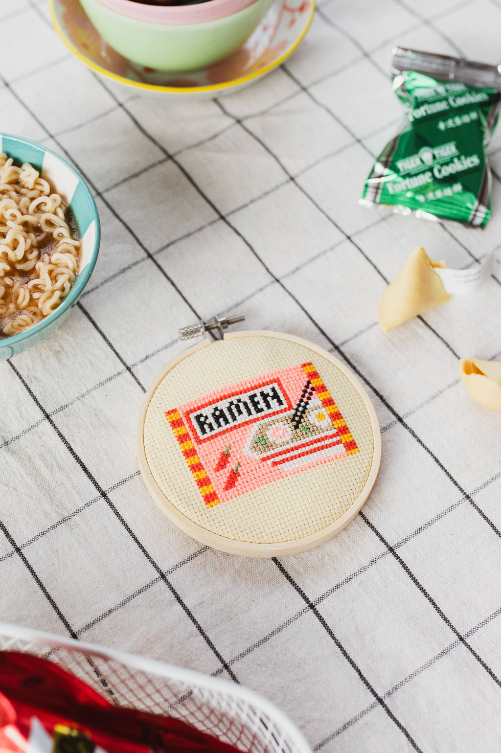 Embroidery hoop with 'RAMEN' design on a tiled floor with noodles and fortune cookies.