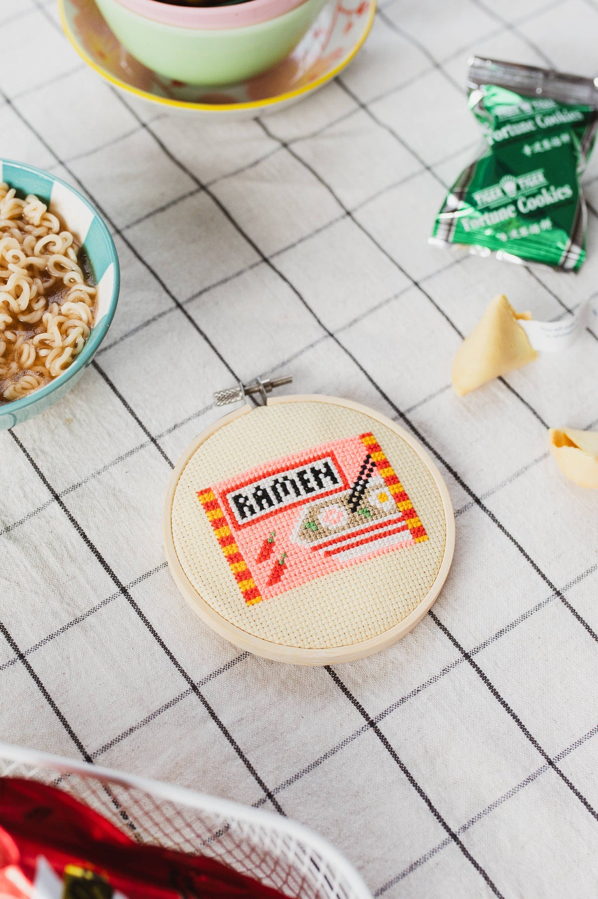 Embroidery hoop with 'RAMEN' design on a tiled floor with noodles and fortune cookies.