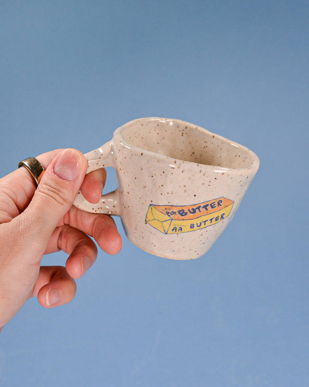Hand holding a ceramic mug with 'Butter' text against a blue background
