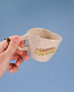 Hand holding a ceramic mug with 'Butter' text against a blue background