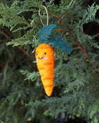 A fuzzy orange carrot Christmas ornament with a smiling face and green leafy top, hanging from a green evergreen tree branch.
