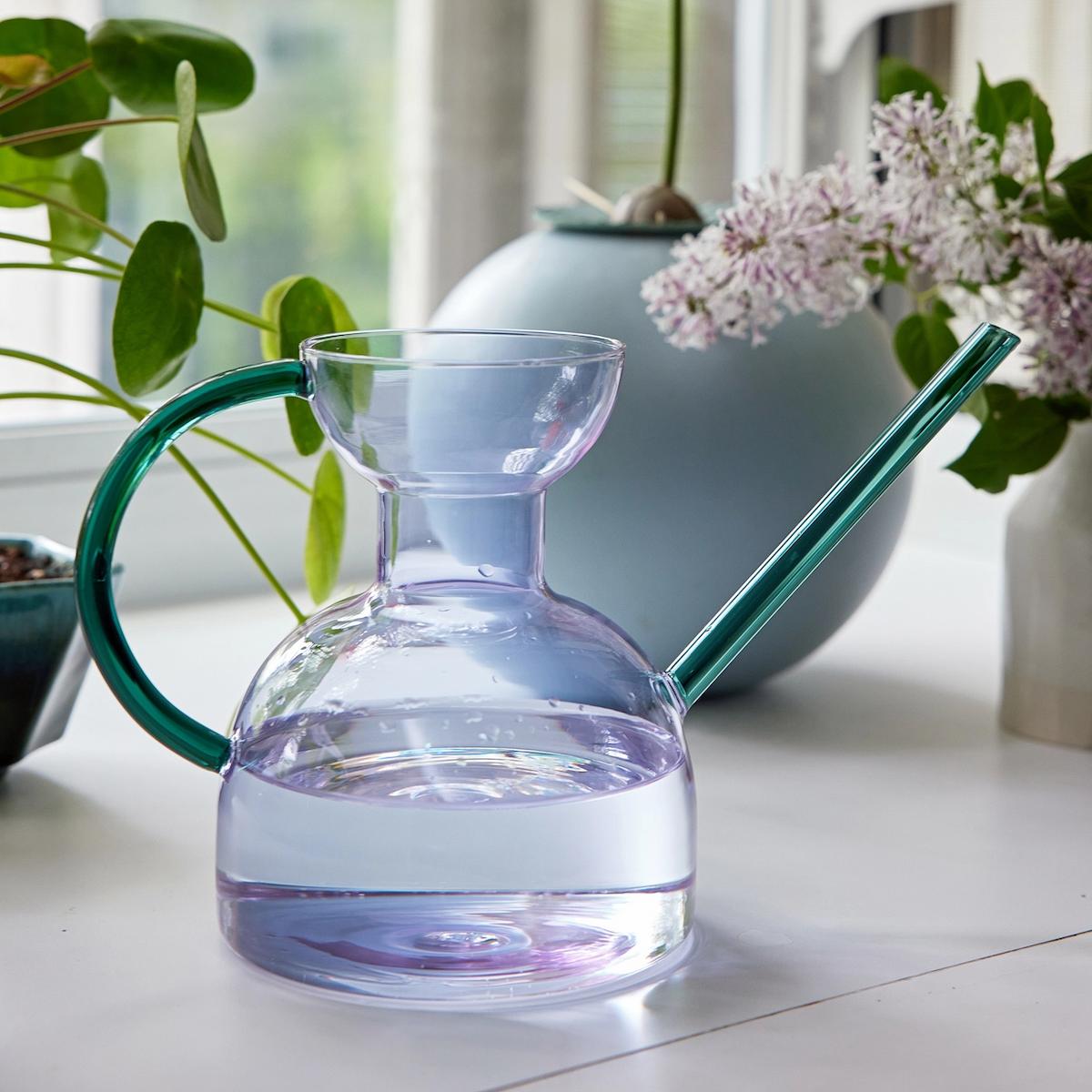Clear glass watering can with green handle on a white surface with plants in the background