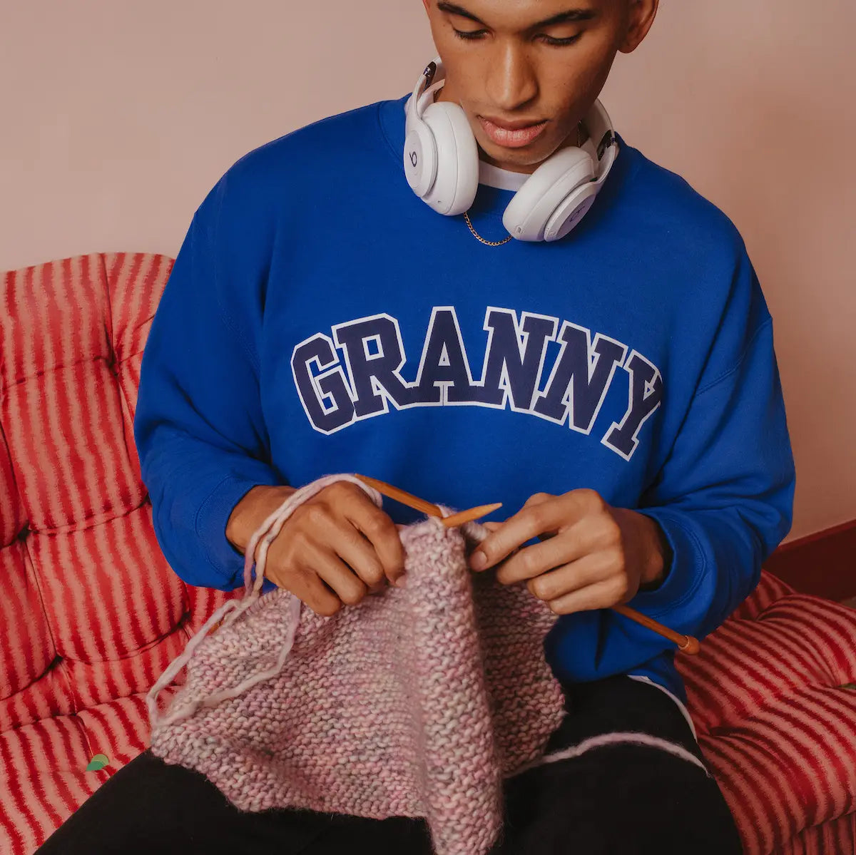 Person wearing a blue sweatshirt with 'GRANNY' text, sitting on a red checkered chair, knitting.