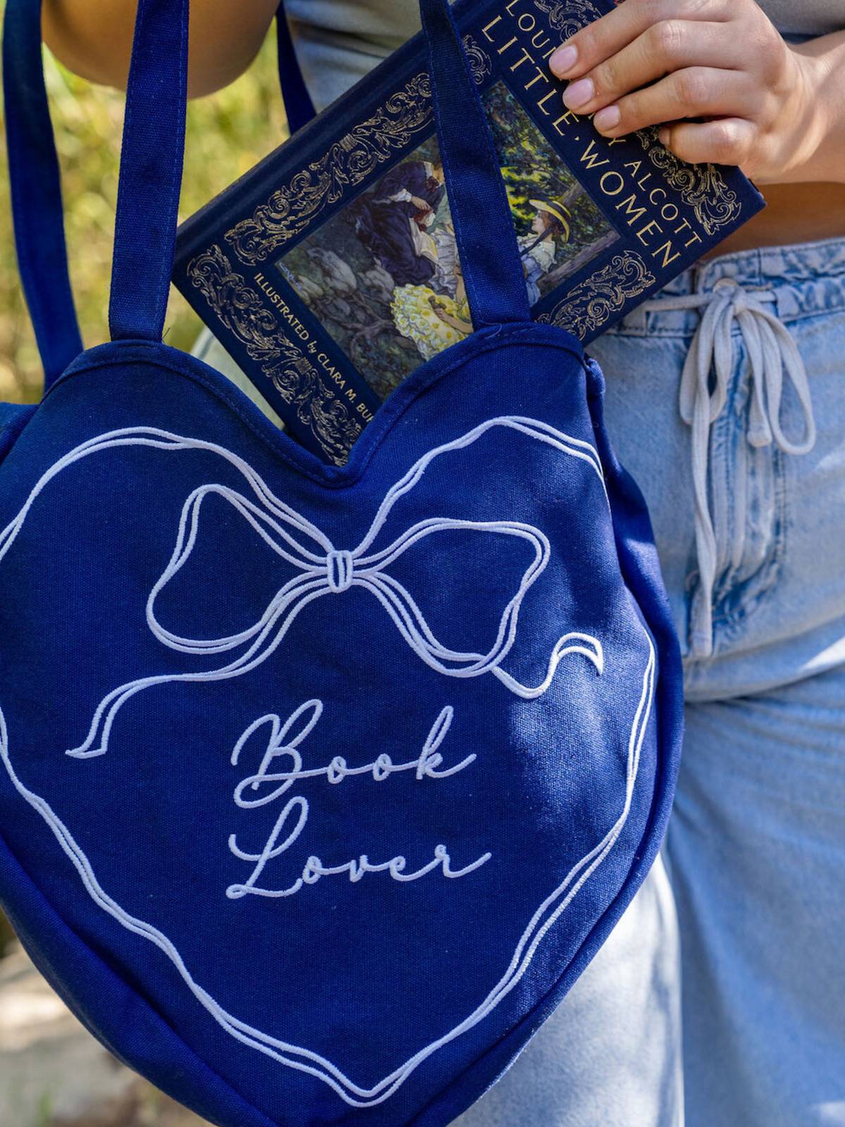 Blue heart-shaped bag with 'Book Lover' text held by a person wearing denim shorts.