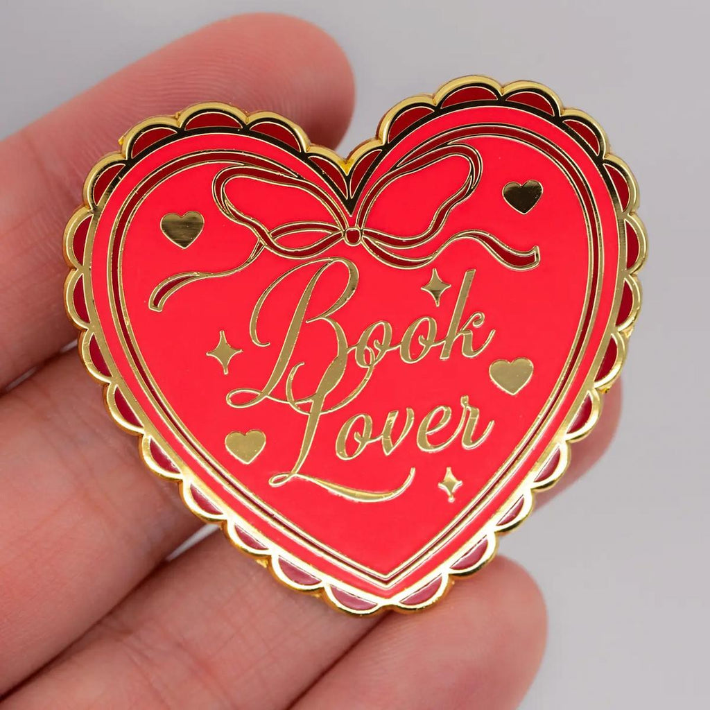 Heart-shaped 'Book Lover' pin held in a hand against a neutral background