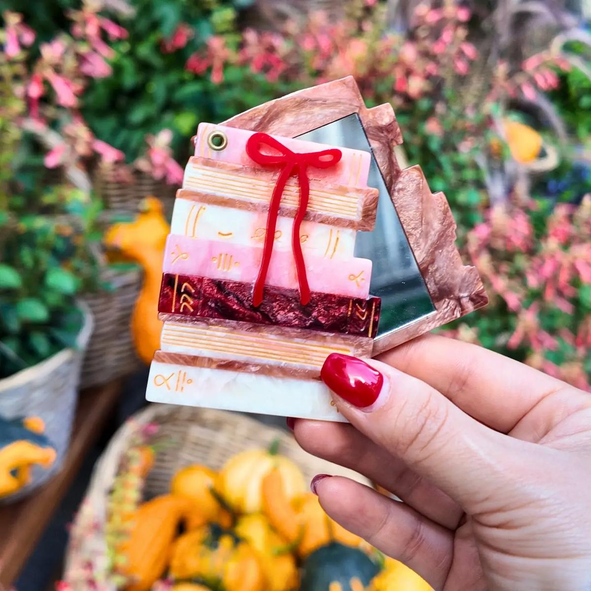 Hand holding a small, decorative mirror with a red bow against a blurred floral background