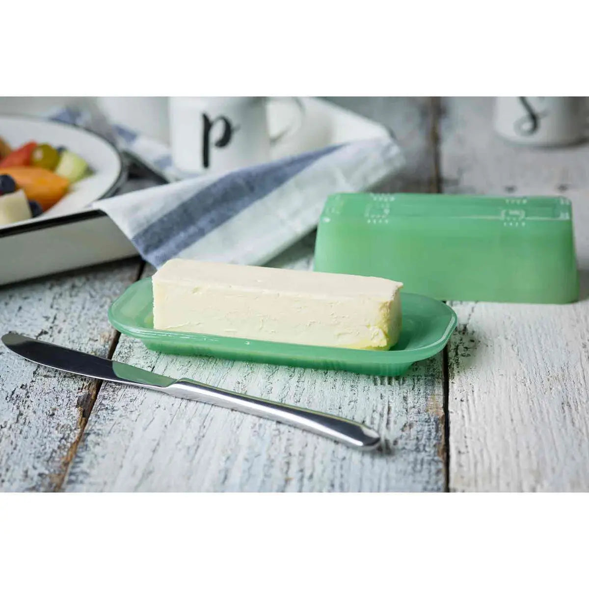 Block of butter on a green plate with a knife, next to a green container, on a wooden surface.