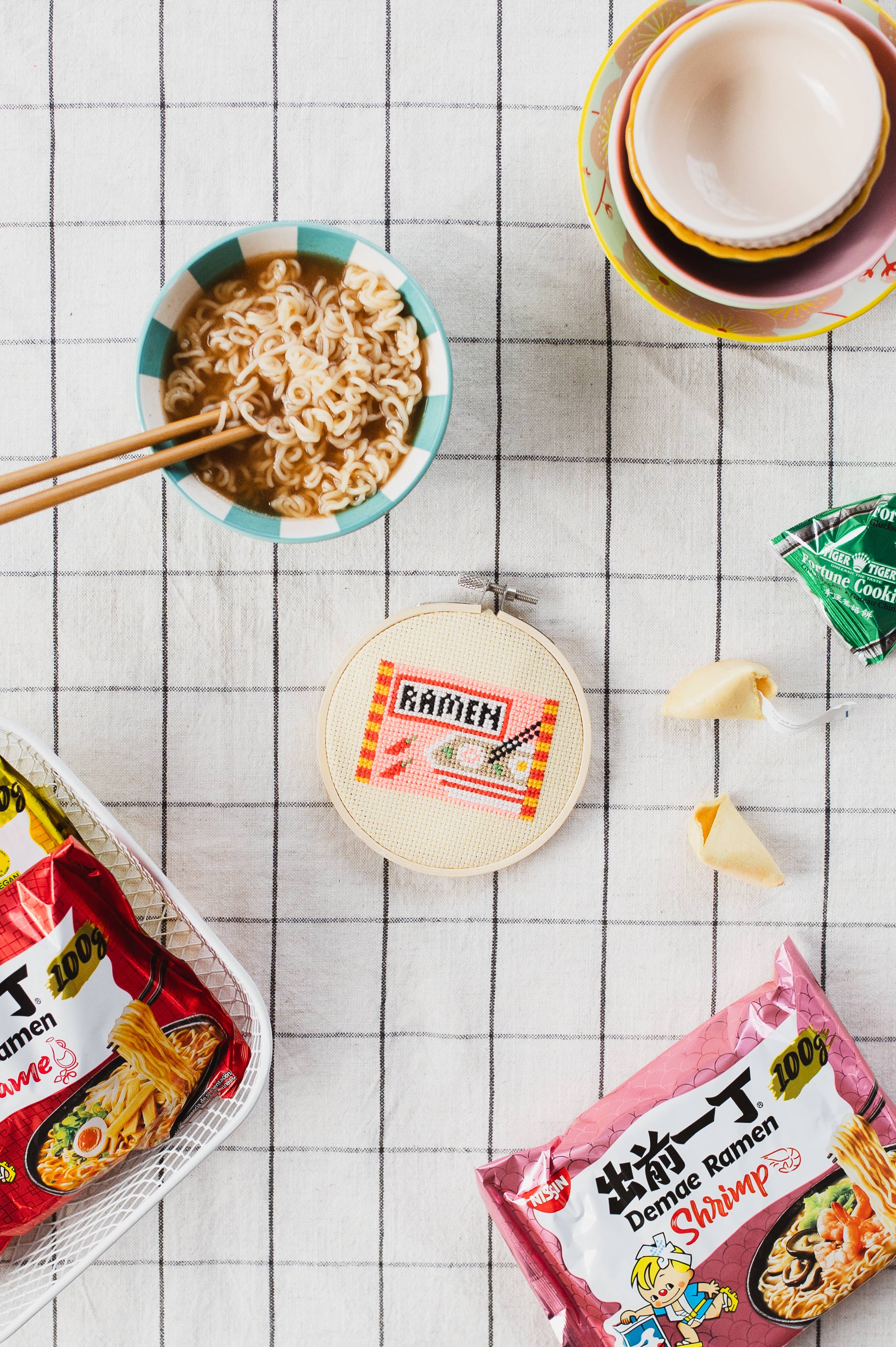 Noodle packages, a bowl of noodles, and a cross stitch ramen design on a checkered tablecloth.