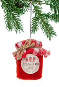 A red felt Christmas ornament shaped like a strawberry jam jar, hanging from a green pine tree branch. The jar has a red and white gingham checkered fabric lid tied with twine, a white round label with two strawberries and the words 'STRAWBERRY JAM', and a silver hanging loop.