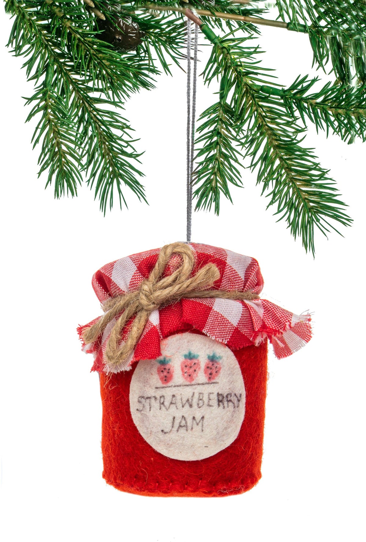 A red felt Christmas ornament shaped like a strawberry jam jar, hanging from a green pine tree branch. The jar has a red and white gingham checkered fabric lid tied with twine, a white round label with two strawberries and the words 'STRAWBERRY JAM', and a silver hanging loop.