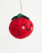 A red felt tomato ornament with a green stem, embroidered black smiley face, and white cheeks, hanging by a thin string against a plain white background.