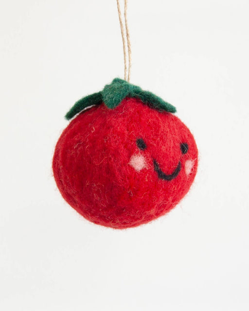 A red felt tomato ornament with a green stem, embroidered black smiley face, and white cheeks, hanging by a thin string against a plain white background.