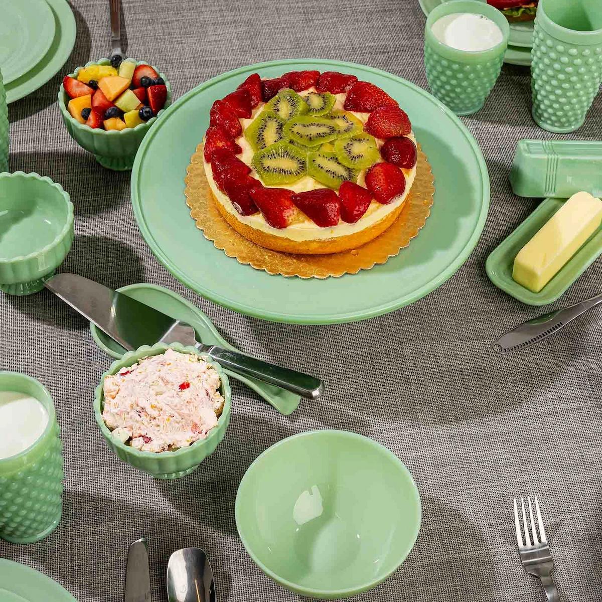 Green ceramic tableware set with a fruit tart and various dishes on a gray tablecloth.