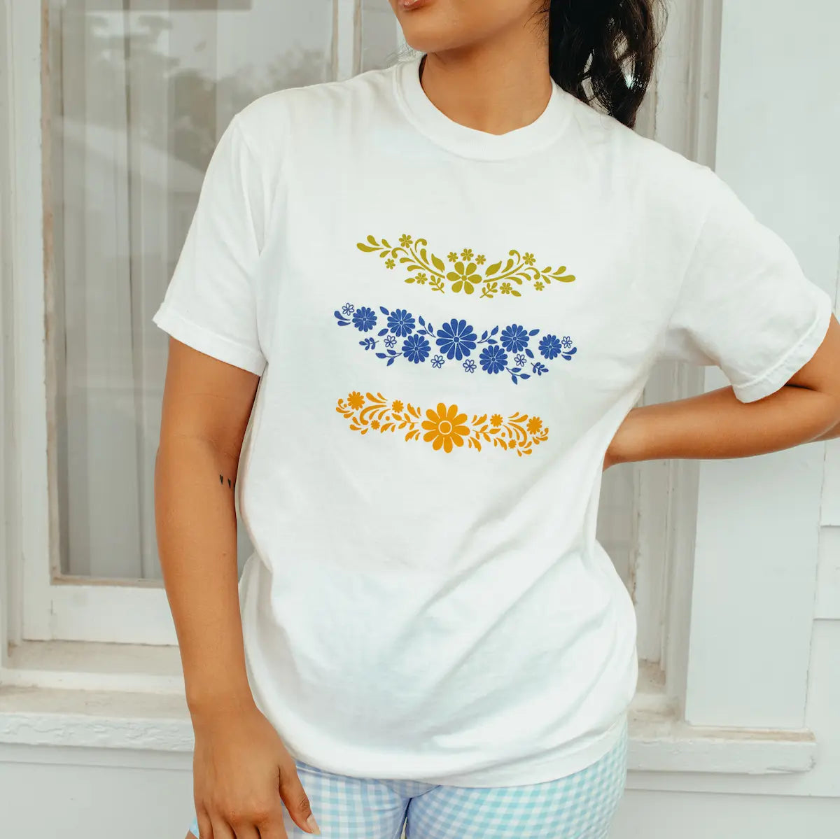 Woman wearing a white t-shirt with Pyrex designs in blue and yellow, standing in front of a white window.