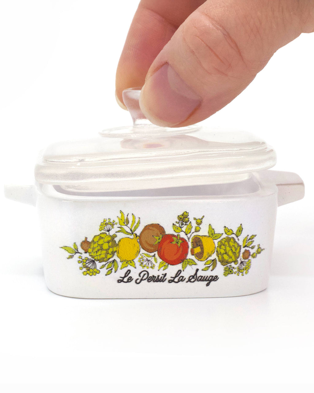 A hand is lifting the clear glass lid of a white ceramic Dutch oven inspired trinket box with a colorful fruit illustration and the brand name 'Le Petit La Bouche' printed on the front, set against a plain white background.