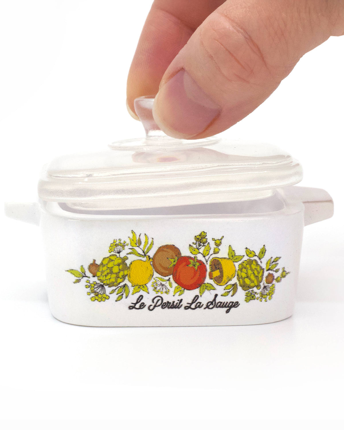 A hand is lifting the clear glass lid of a white ceramic Dutch oven inspired trinket box with a colorful fruit illustration and the brand name 'Le Petit La Bouche' printed on the front, set against a plain white background.