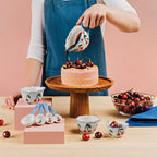 Person decorating a cake with cherries using cherry-themed kitchenware on a pink background