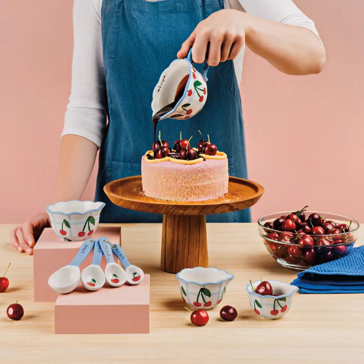 Person decorating a cake with cherries using cherry-themed kitchenware on a pink background