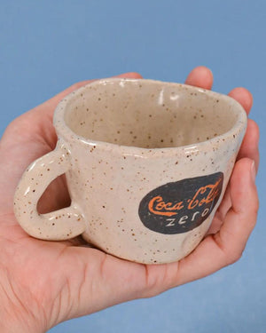 Speckled ceramic mug with Coca-Cola Zero logo held in a hand against a blue background