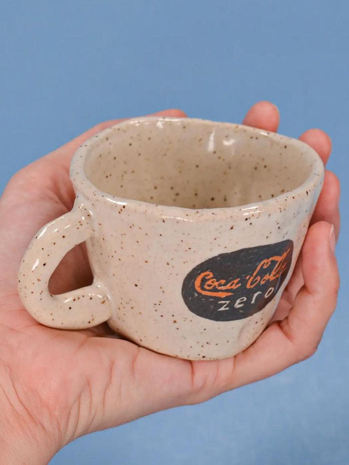 Speckled ceramic mug with Coca-Cola Zero logo held in a hand against a blue background