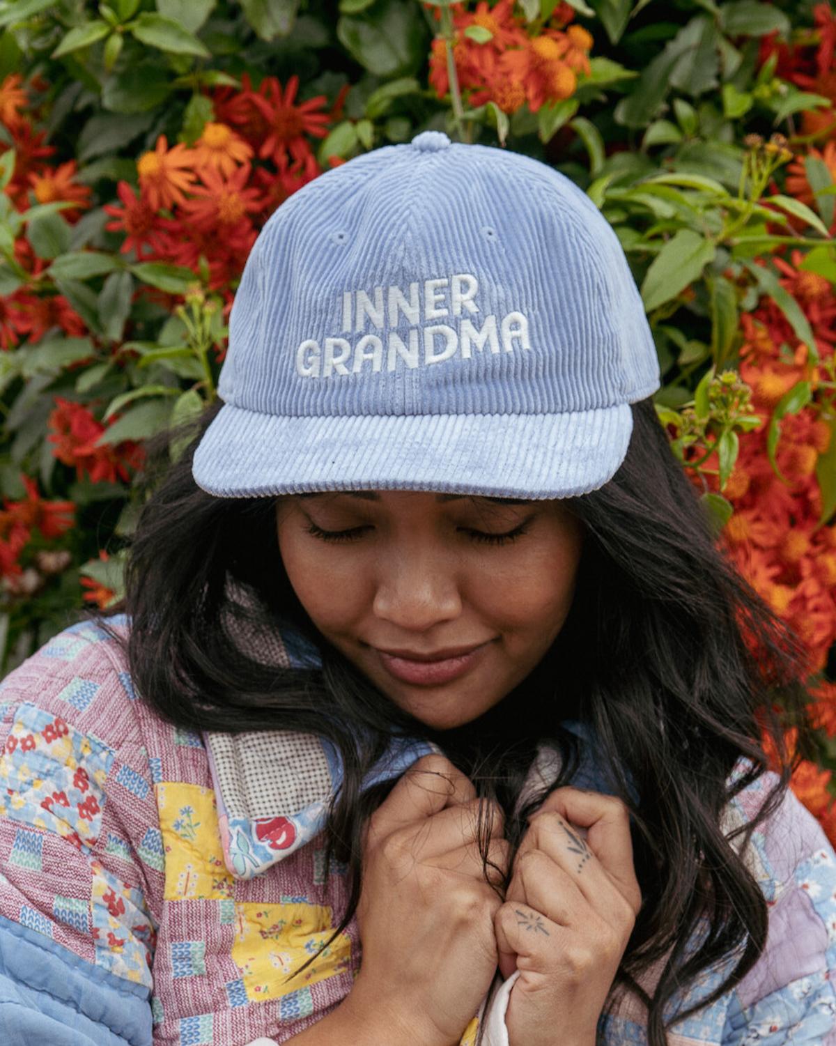 Person wearing a blue cap with 'INNER GRANDMA' text, standing in front of a floral background.