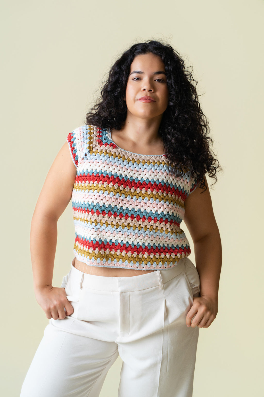 Person wearing a colorful crochet cap sleeve top and white pants against a beige background