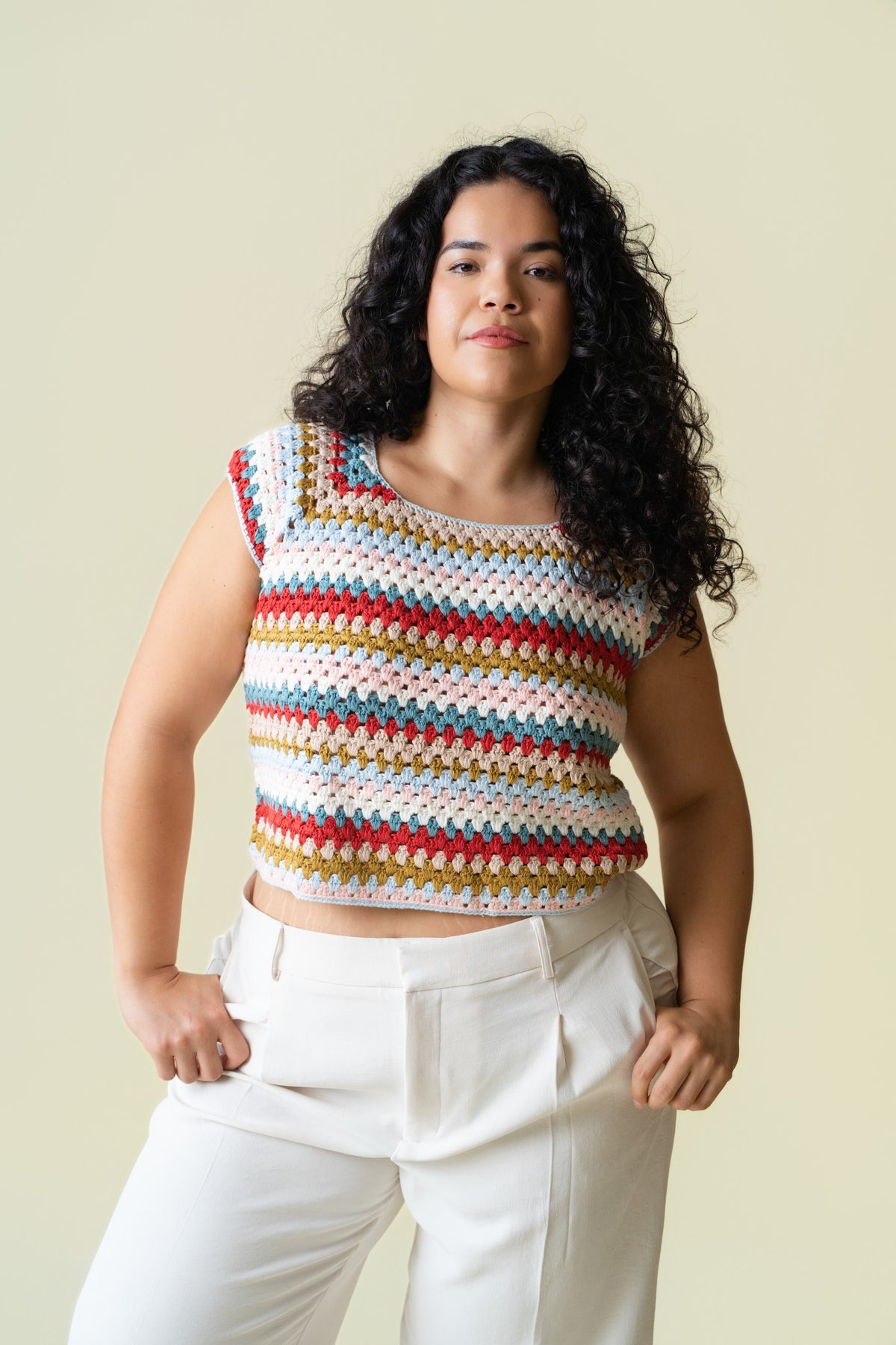 Person wearing a colorful crochet cap sleeve top and white pants against a beige background