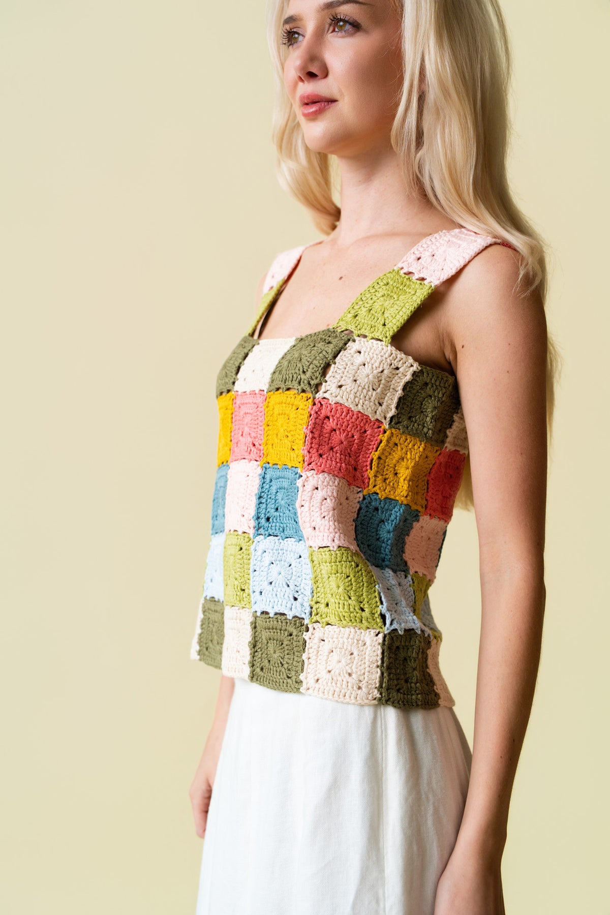 Side of person wearing a colorful crocheted tank top and white skirt against a beige background