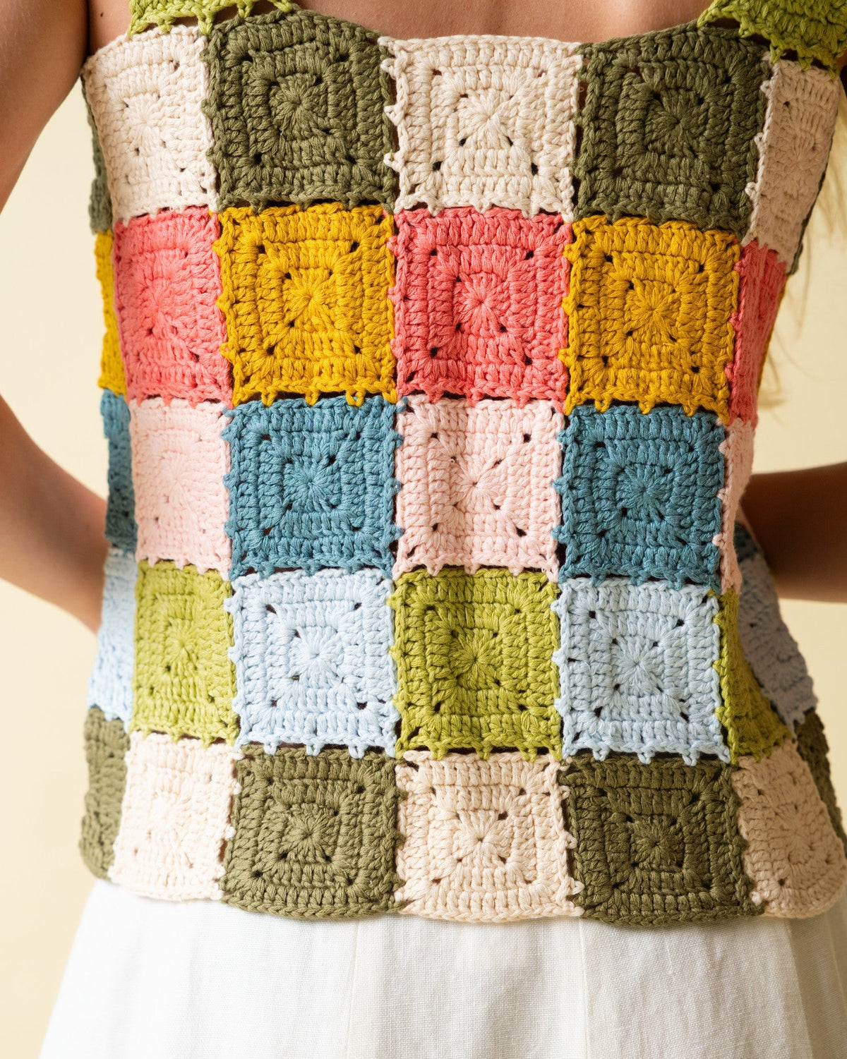 Back of person wearing a colorful crocheted square pattern top and white skirt against a neutral background