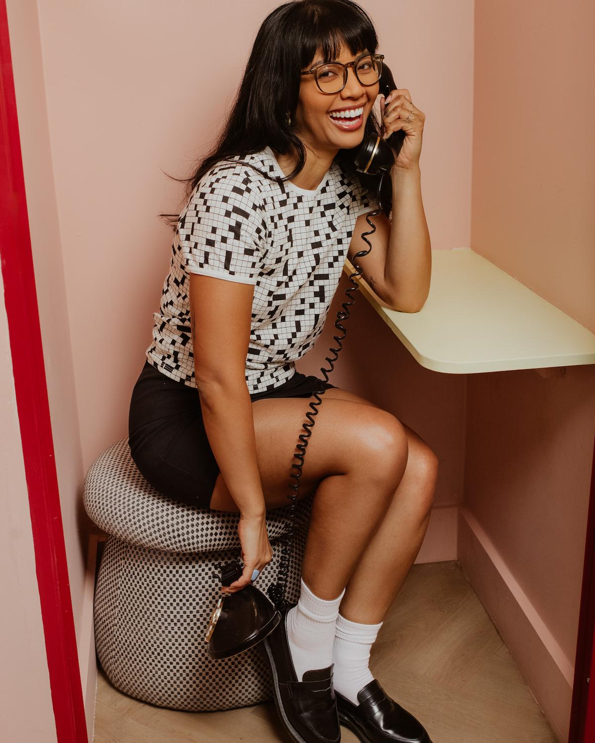 Woman sitting on a stool in a booth, talking on a vintage phone with a pink wall background.