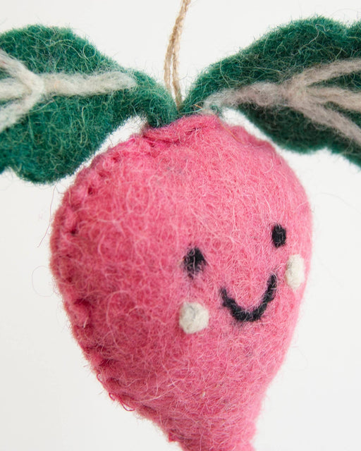 A close-up of a handmade pink felt radish ornament with green felt leaves and a smiling embroidered face, hanging against a plain white background.