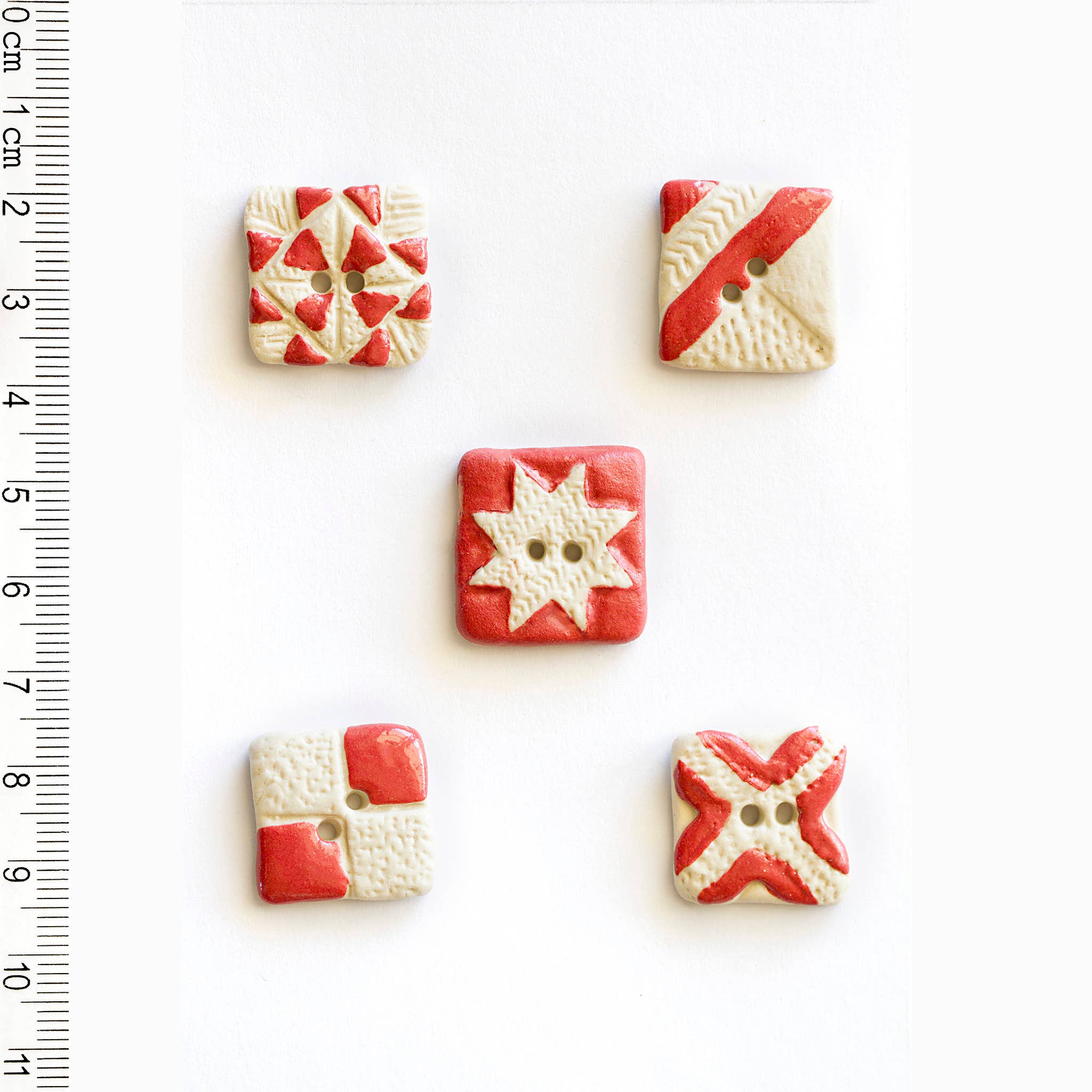 Five decorative buttons with red and white patterns on a white background, accompanied by a ruler for scale.
