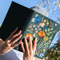 Person holding a black book with floral embroidery against a blue sky.
