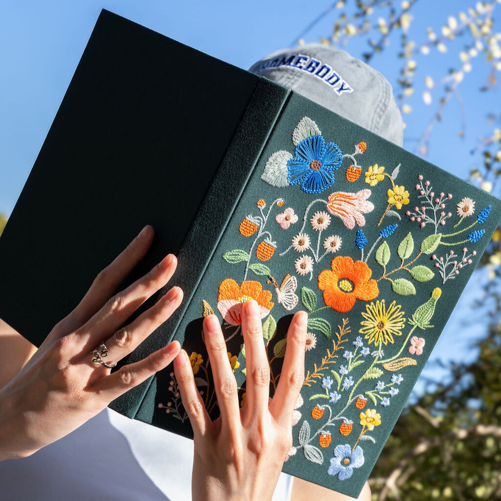 Person holding a black book with floral embroidery against a blue sky.