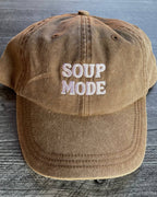 A brown washed cotton baseball cap with 'SOUP MODE' embroidered in white on the front, displayed on a gray wood-textured background.