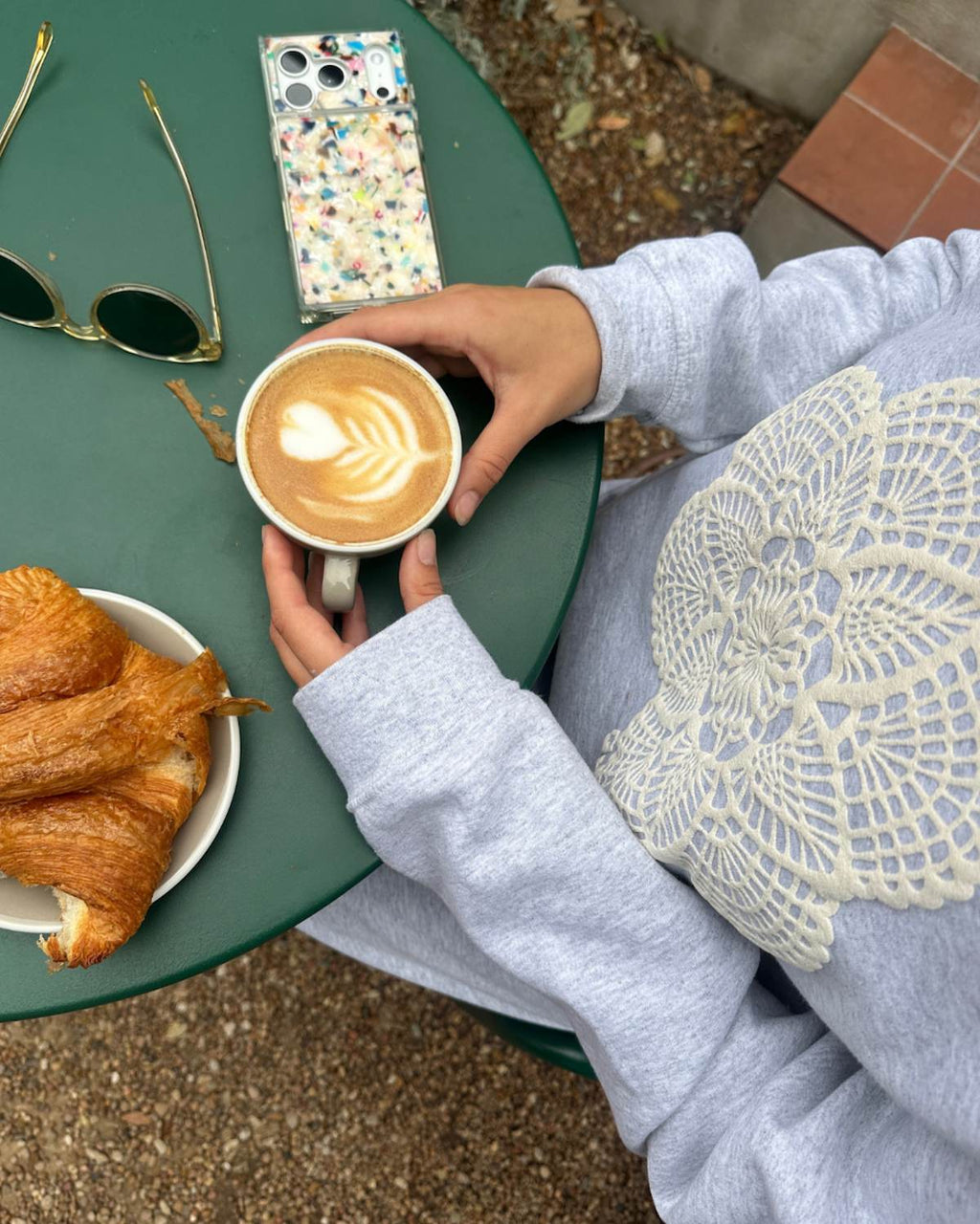 Person holding a cup of coffee with a croissant on a green table outdoors.
