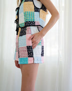 Person wearing a sleeveless patchwork quilted vest and matching patchwork quilted shorts, standing in front of white curtains.