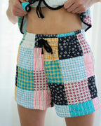A person wearing patchwork patchwork shorts with a mix of gingham, striped, and floral fabric panels in pink, blue, yellow, black, and white, paired with a matching tie-front crop top.
