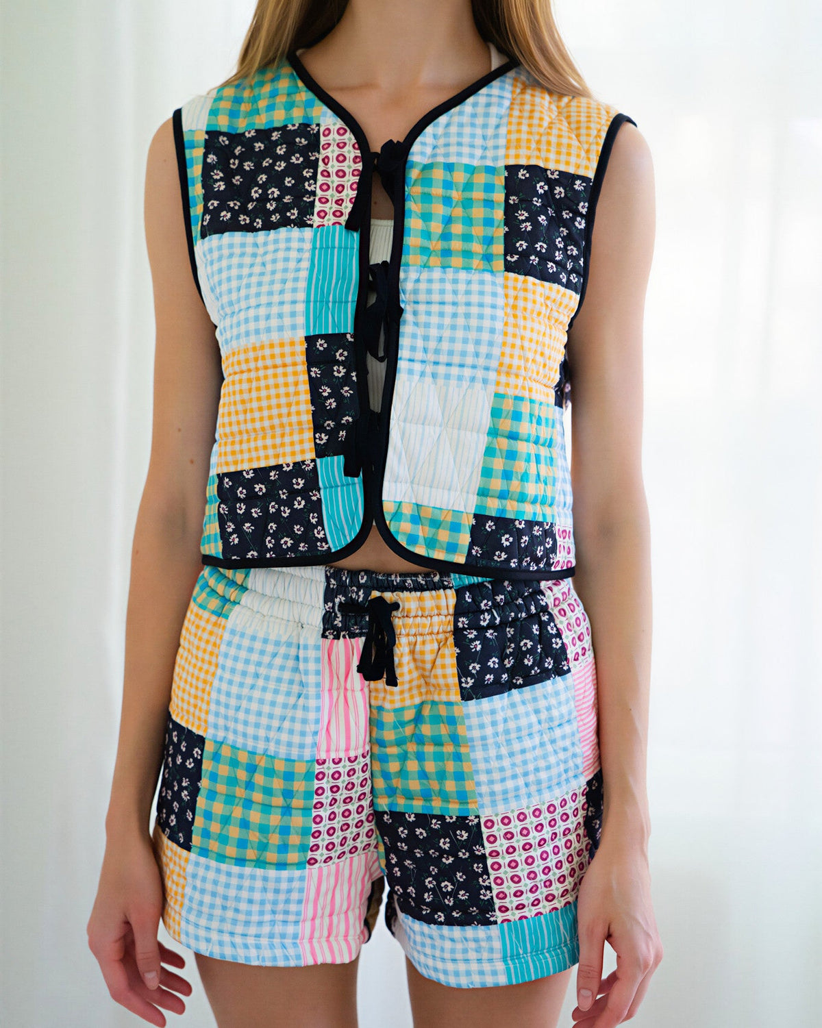 Person wearing a matching sleeveless vest and shorts set with a colorful patchwork print of gingham, floral, and solid squares in yellow, blue, pink, black, and white, photographed against a bright white curtain background.