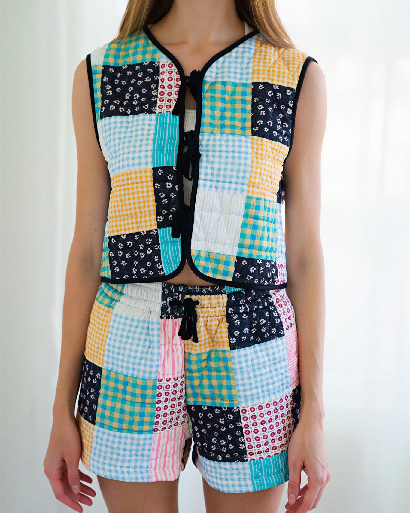 Person wearing a matching sleeveless vest and shorts set with a colorful patchwork print of gingham, floral, and solid squares in yellow, blue, pink, black, and white, photographed against a bright white curtain background.