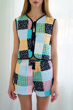 Person wearing a matching sleeveless vest and shorts set with a colorful patchwork print of gingham, floral, and solid squares in yellow, blue, pink, black, and white, photographed against a bright white curtain background.