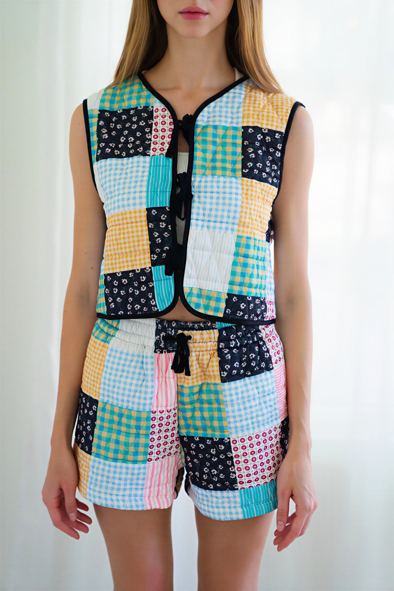 Person wearing a matching sleeveless vest and shorts set with a colorful patchwork print of gingham, floral, and solid squares in yellow, blue, pink, black, and white, photographed against a bright white curtain background.