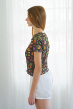 A person in profile wearing a short-sleeve crewneck t-shirt with a colorful vegetable print, paired with white shorts, standing in front of a white curtain.