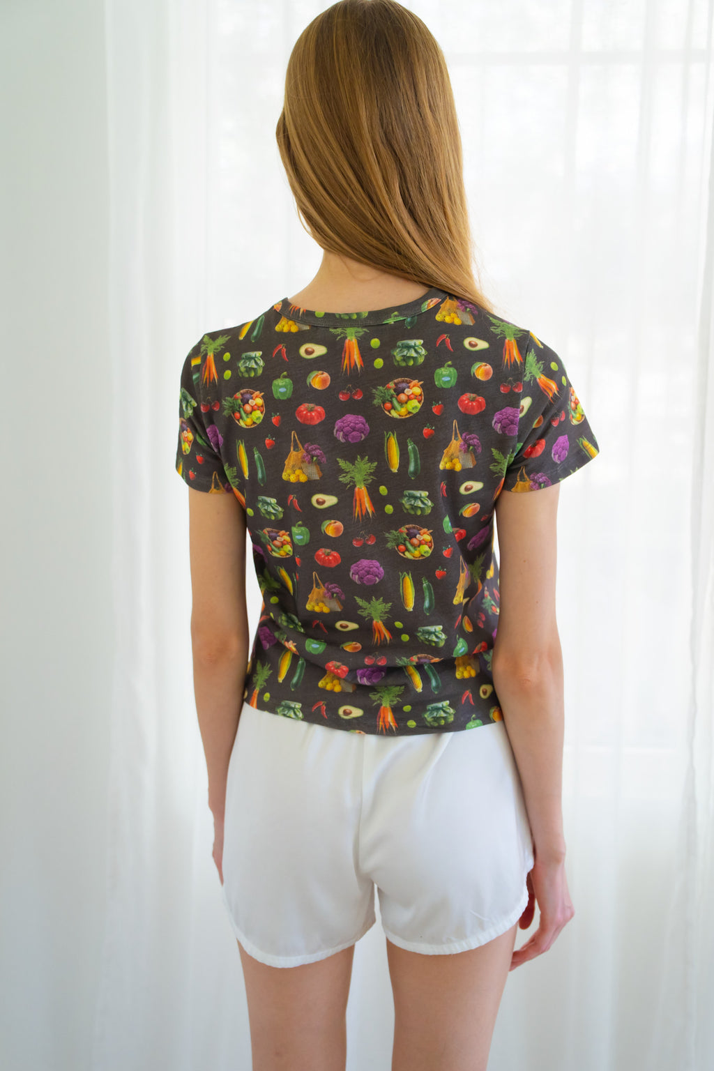 Back of person wearing a dark charcoal gray t-shirt with a colorful vegetable print and white shorts, standing in front of a white curtain.