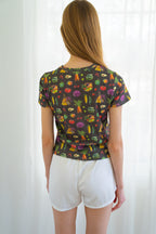 Back of person wearing a dark charcoal gray t-shirt with a colorful vegetable print and white shorts, standing in front of a white curtain.